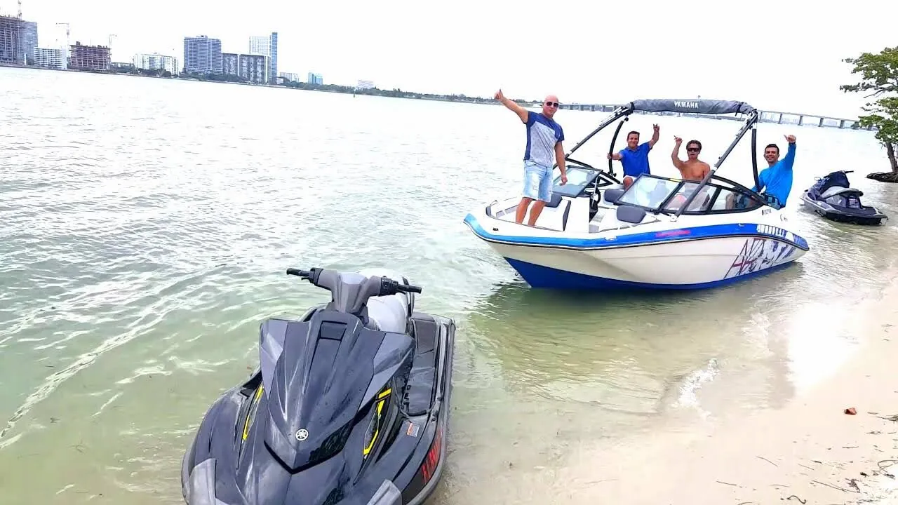 Boat and jet ski pulled up to a sandy island beach with guests aboard and Miami skyline in the distance