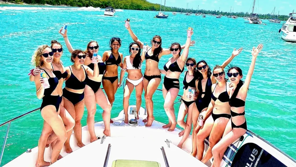 Bachelorette party of women in black swimsuits cheering on the bow of a boat in Miami's turquoise water